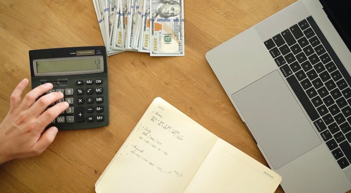 Person calculating Europe travel budget with calculator, cash and laptop on desk, planning trip expenses  